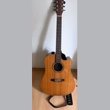 Akustická kytara s cutawayem v přirozené dřevěné barvě. Na hlavě hmatníku je viditelné logo Cort. Nástroj je umístěn v černém stojanu a má připojený…