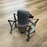 Stříbrný kondenzátorový mikrofon s kovovou síťkou, umístěný v šedém pavoukovém držáku. Na spodní části těla je viditelné červené logo s nápisem AKG a…
