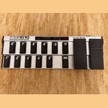 Stříbrný MIDI pedál s černými spínači a dvěma expresními pedály. Na levé straně je nápis FCB1010 MIDI FOOT CONTROLLER a nad spínači jsou sekce PRESET…