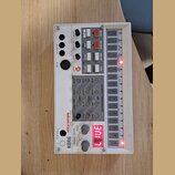 Bílý sampler s názvy KORG a volca sample DIGITAL SAMPLE LESS SEQUENCER. Zařízení obsahuje řadu posuvníků, tlačítek a červeně svítící displej s nápise…