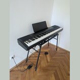 Černý digitální klavír umístěný na černém kovovém stojanu. Nástroj má bílé a černé klávesy, nad nimi se nachází ovládací panel s tlačítky a černý not…
