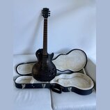 Černá elektrická kytara s tmavým dřevěným povrchem a v černé hardshell case s bílou vnitřní výplní. Na hlavě nástroje je zlatý nápis Gibson. Kytara m…