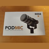 Fotografie znázorňuje bílé originální balení mikrofonu. Na krabici je zobrazen černý dynamický mikrofon s kovovou mřížkou a nápisy RØDE, PODMIC a DYN…