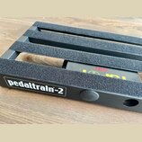 Černý kovový pedalboard s nápisem pedaltrain-2 v bílém rámečku. Horní lišty jsou potaženy šedým protiskluzovým materiálem. Pod deskou je vidět část č…
