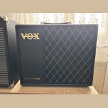 Černý kytarový kombozesilovač s pravou stranou pokrytou černou tkaninou s bílým kosočtvercovým vzorem. V horní levé části je umístěno zlaté logo VOX…