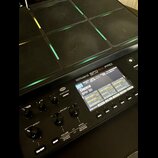 Černý sampling pad značky Roland s nápisem SPD-SX PRO. Zařízení má mřížku gumových padů s podsvícením v různých barvách. Displej zobrazuje název „001…