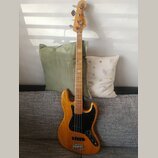Elektrická basová kytara s přírodním dřevěným povrchem těla a hmatníkem. Na hlavě je vidět nápis Fender Jazz Bass. Instrument má černý pickguard a čt…