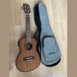 Hnědý dřevěný ukulele s doprovodným šedým textilem v podobě obalu. Na hlavě nástroje i na přední straně obalu je bílý nápis CASCHA. Na hmatníku jsou…