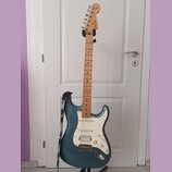 Elektrická kytara s modrým kovovým povrchem těla a světle hnědým javorovým hmatníkem. Na hlavě je viditelné logo Fender. Kytara má bílý pickguard, tř…