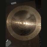 Kruhový činel z mosazného kovu s výrazným žebrováním v centrální části a kováními (důlky) po celém okraji. Na povrchu je černé logo Paiste a nad ním…