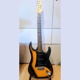 Elektrická kytara s trupem v barevně sunburst (černá a žlutá) a černým pickguardem. Na hlavě hmatníku je viditelný nápis „G&L“ a pod ním text „by Leo…