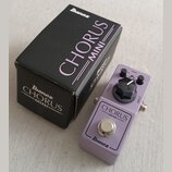 Fialový efekt pedál s nápisy CHORUS MINI a Ibanez. Na horní části je jeden černý ovládací knoflík a konektory OUTPUT a INPUT. Vedle pedálu je černá k…
