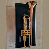 Zlatý mosazný trumpet položený na černém kufru. Na kufru je bílý nápis YAMAHA v obdélníkovém rámečku. Nástroj vykazuje známky používání v podobě drob…