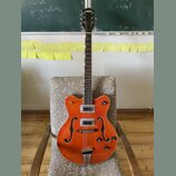 Oranžová polodupatá elektrická kytara s hnědým hmatníkem a bílým lemováním. Na hlavě je bílý nápis GRETSCH a na spodní části korpusu je kovový most s…