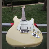 Světle žlutá elektrická kytara s bílým pickguardem a hnedým hmatníkem. Nástroj má tři snímače typu single-coil, jeden tremolo páku a tři bílé oválné…