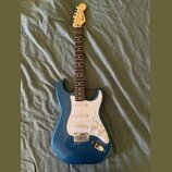 Modrá elektrická kytara s bílým pickguardem a světle hnědým hmatníkem. Na hlavě je viditelné logo Fender. Nástroj má tři snímače, bílé ovládací knofl…