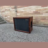 Hnědý basový reproduktor v dřevěném korpusu s černou textilní maskou. V pravém horním rohu masky je umístěn štítek s nápisem „gbr Custom Cabinets“. P…
