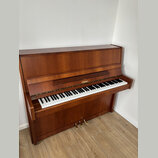 Hnědé dřevěné piano s otevřenou klaviaturou. Na přihradě nad klávesami je zlatý nápis „Behringer“. Nástroj vykazuje známky běžného používání, povrch…