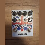 Bílý kytarový efekt s červenými a modrými grafickými prvky. Na zařízení jsou nápisy Plexi-Drive Deluxe a WAMPLER BUILT IN USA. Horní řada ovládacích…