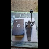 Na fotografii je černý obdélníkový box s nápisem SnareBox a logem MEINL v bílém kruhu. Vedle něj je šedý basový pedál s logem ludwig. Oba předměty le…