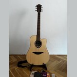 Akustická kytara s vyřezem v korpusu, světlým přírodním povrchem lícové desky a tmavým hmatníkem. Na hlavě je viditelné logo LG. Kytara stojí na čern…