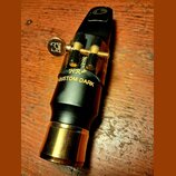 Černý saxofonový mouthpiece s mosazným prstencem u patky. Na povrchu je zlatým písmem nápis HR* CUSTOM DARK a nad ním nečitelný podpis. Mundstuk je v…