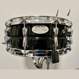 Černý buben s chromovaným obručováním a mechanismy. Na korpusu je oválný štítek s nápisy CONCERT, YAMAHA a JAPAN. Povrch nástroje je glossy, v odrazu…