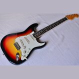 Elektrická kytara s trupem v barevnosti sunburst a bílým pickguardem. Na hmatníku jsou vidět čtyři snímače s bílými kryty a tři ovládací knoflíky, z…