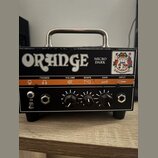 Černý kytarový zesilovač s kovovou rukojetí a oranžovým pruhem na předním panelu. Na horní části je bílý nápis ORANGE a vedle něj text MICRO DARK, v…