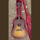 Akustická kytara s korpusem v barevném přechodu od žluté k tmavě hnědé (sunburst) a tmavým hmatníkem. Na hlavě nástroje je bílý nápis Gibson. Kytara…