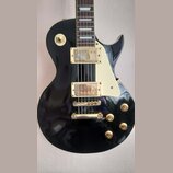 Černá elektrická kytara s typickým tvarem singlecut a krémovým pickguardem. Má dva humbuckery se zlatými kryty, zlatý kobylník a čtyři zlaté ovládací…