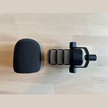 Na fotografii je černý mikrofon v držáku a samostatná černá pěnová clona. Mikrofon má kovovou mřížku s viditelnými stříbrnými prvky. Na zařízení nejs…