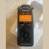 Černý digitální záznamový zařízení s nápisem TASCAM a označením modelu DR-05. Displej je zapnutý s oranžovým podsvícením a zobrazeným textem 00H00M00…