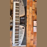 Stříbro-černý digitální klavír s bílými a černými klávesami. Na přístroji jsou viditelný nápisy Roland a E-60 MUSIC WORKSTATION. Povrch vykazuje znám…