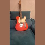 Elektrická kytara s červeným trupem, bílým pickguardem a hnedým hmatníkem. Na hlavě nástroje je viditelné logo Fender. Povrch instrumentu vypadá čist…