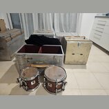 Na fotografii jsou dva hluboké bubny s tmavým dřevěným povrchem a kovovými obručemi. Vedle nich jsou čtyři přepravní kufry z kovu a světlého materiál…