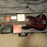 V otevřeném kufru leží elektrická basová kytara s tmavě hnědým sunburst povrchem a hnědým hmatníkem. Na těle je vidět tmavě červený pickguard a kový…