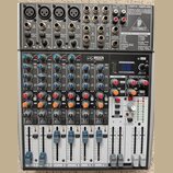 Šedý mixážní pult značky BEHRINGER s modelem XENYX X2022USB. Zařízení obsahuje několik vstupních kanálů s regulačními knoflíky, posuvníky hlasitosti…