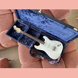 Bílá elektrická kytara typu Stratocaster v otevřeném tvrdém kufru s modrým polstrováním. Na hlavě hmatníku je viditelné logo Fender. Kytara má tři sn…