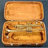 Zlatý trumpet v hnědém tvrdém kufru s plišovou výstlkou. Nástroj je v lesklém povrchu, bez viditelných značek či nápisů. Povrch nástroje i vnitřek ku…