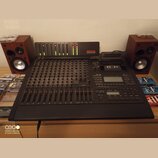 Černý digitální mixážní pult s řadou posuvníků a ovládacích knoflíků. V horní části je umístěn modul s LED indikátory a nápisem TASCAU DIGITAL STUDIO…