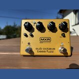 Zlatý efekt pedál s nápisy MXR a BASS INNOVATIONS. Na horní straně jsou čtyři černé oválné knoflíky s popisky GAIN, FUZZ, OCTAVE a DRY. Ve spodní čás…