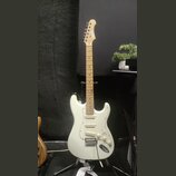 Bílá elektrická kytara typu Stratocaster s jasným dřevěným hmatníkem a hlavou. Na hlavě je viditelné logo Squier. Nástroj má tři snímače typu single-…