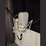 Stříbrný kondenzátorový mikrofon v šokabsorbujícím držáku. Na těle mikrofonu je viditelný nápis NEUMANN a text MADE IN GERMANY. Povrch mikrofonu i dr…