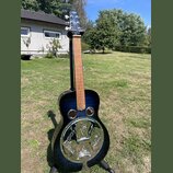 Tmavě modrá akustická kytara typu resonator s kovovým kruhovým ozvučovadlem na přední dece. Na hmatníku jsou vidět bílé polohové značky a na hlavě ná…