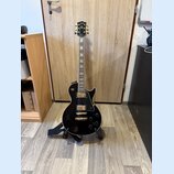 Černá elektrická kytara s gold hardwarem umístěná na stojanu. Na hlavě hmatníku je viditelné logo Epiphone. Kytara má dva humbuckery, čtyři ovládací…