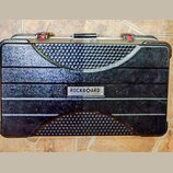Černý transportní kufr s texturovaným povrchem a vodorovnými žebrováním. Uprostřed je umístěn kovový štítek s nápisem ROCKBOARD by WARWICK. Povrch vy…