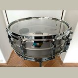Stříbrný snare drum s chromovaným obručováním a mechanismem. Na blaně je viditelný nápis „REMO“ a „HEAVY CLEAR“. Povrch nástroje je hladký, bez vidit…