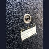 Detail černého povrchu basového komba s texturou. Na povrchu je kovový konektor a štítek s nápisy: „ELECTRONICS CORP“, logo „Peavey“ a adresa „711 A…