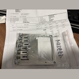 Fotografie zachycuje kovový bridge na elektrickou kytaru v originálním plastovém obalu, který leží na fakturním dokladu. Bridge je stříbrné barvy s š…